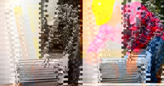 A technician in a yellow hard hat repairs an air conditioner.
