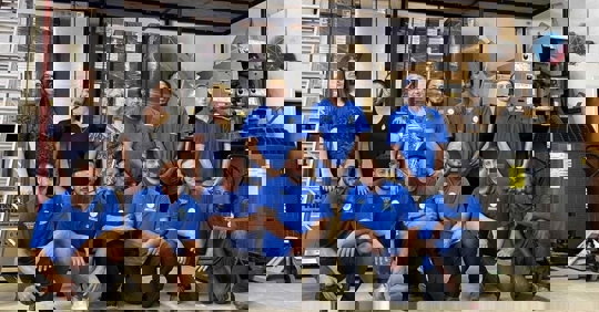 A team of twelve diverse staff pose for a group shot in their warehouse, wearing blue shirts with the company logo.