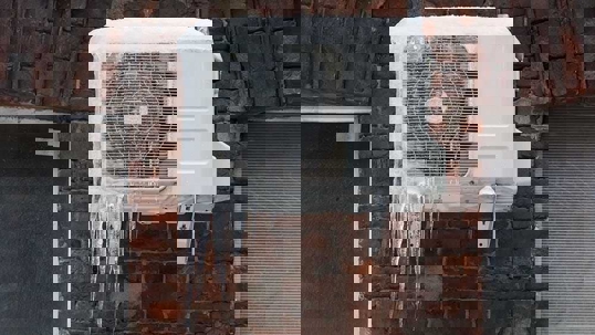 A white air conditioning unit is covered in ice and icicles, attached to a brick wall.